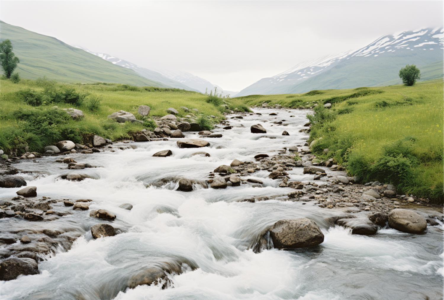 A river running through open green country with snow-capped hills rising behind it